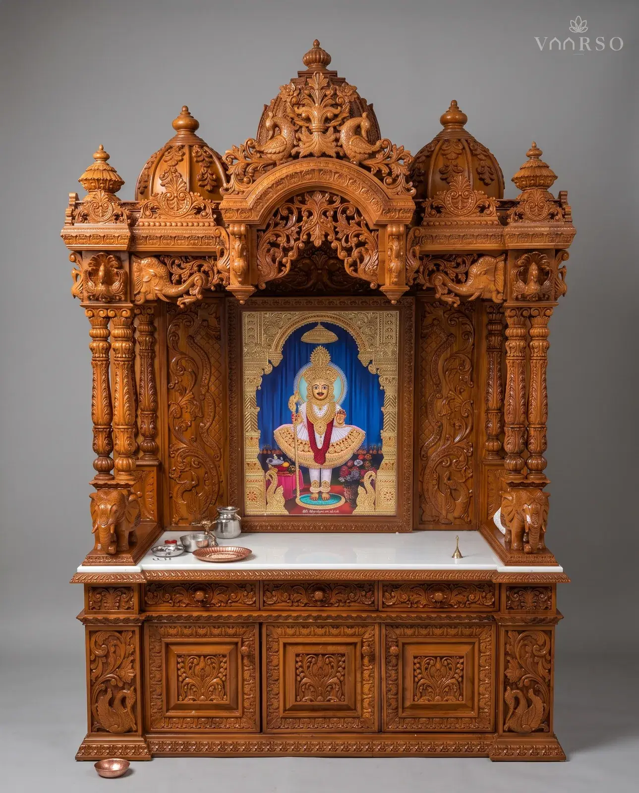A wide mandir in a warm natural polish, a domed crest with peacocks over a carved arch, elephants standing at the columns, a framed picture on a white marble shelf, and drawers over three deeply carved cupboard doors