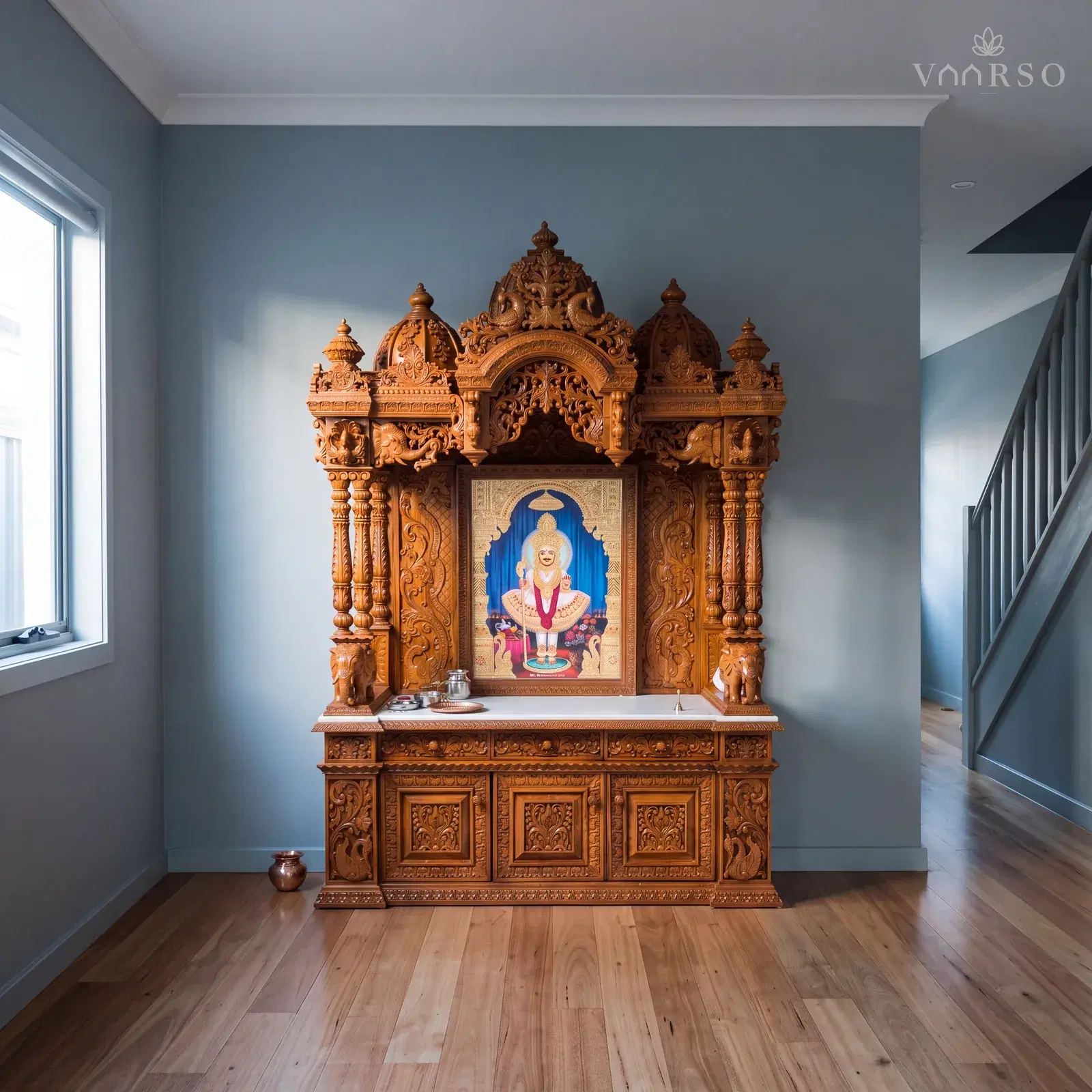 The same mandir in a narrow townhouse living room, the stair rising at the far end, blackbutt boards underfoot and a small brass kalash on the floor beside it