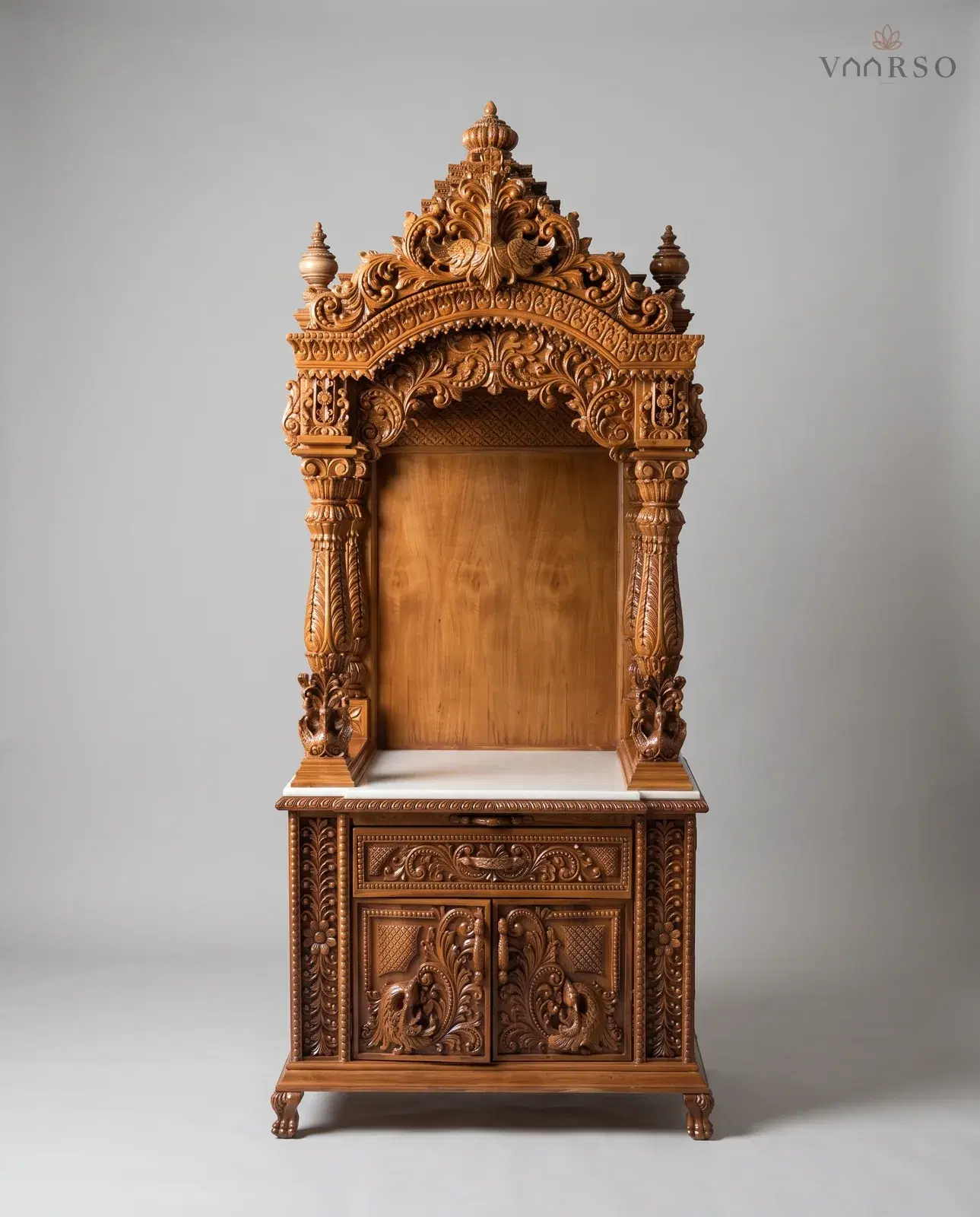 A tall narrow mandir in a warm natural polish, a stepped crest with peacocks over a carved arch, an empty shrine on two carved columns with a white marble shelf, and one drawer over peacock-carved cupboard doors