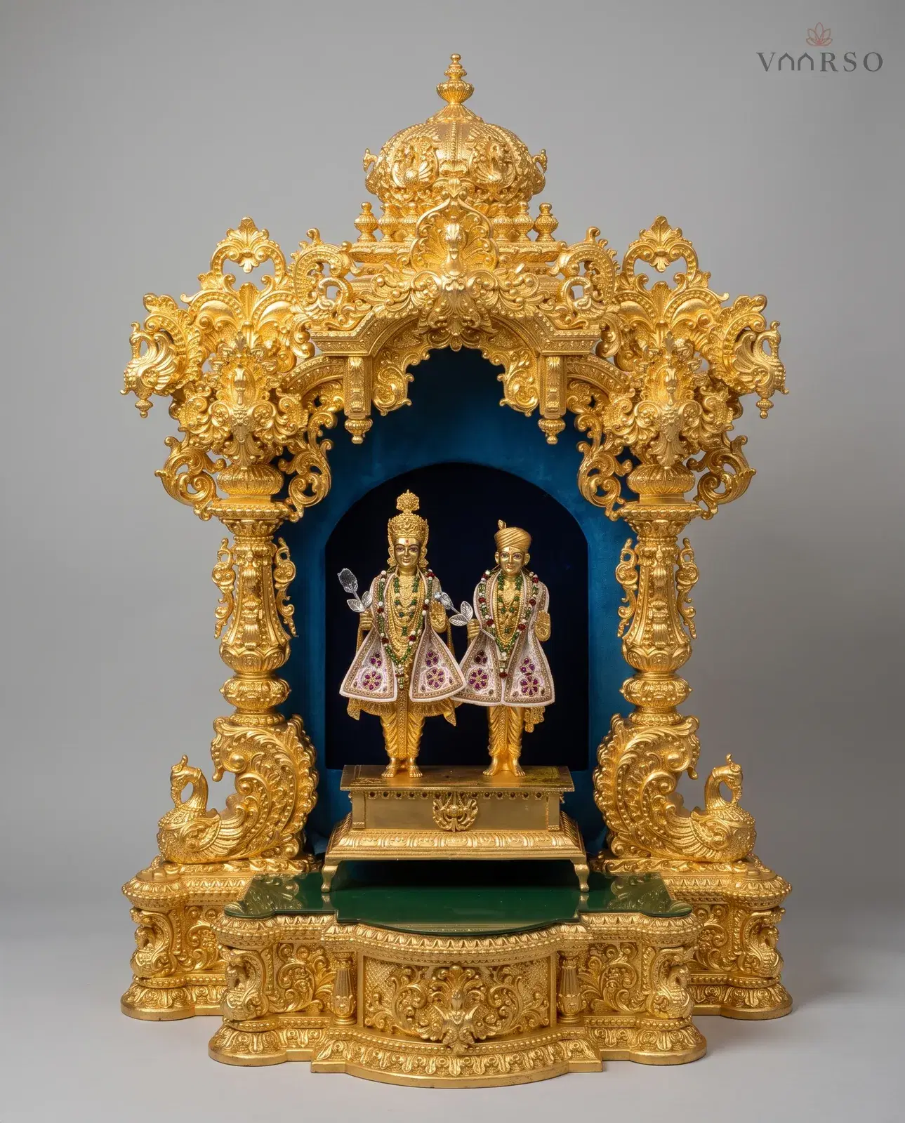 A small gold mandir on a stepped gold plinth, a domed crest and pierced side crests on turned columns, peacocks at the base, and two dressed murtis standing in a deep blue velvet-lined niche