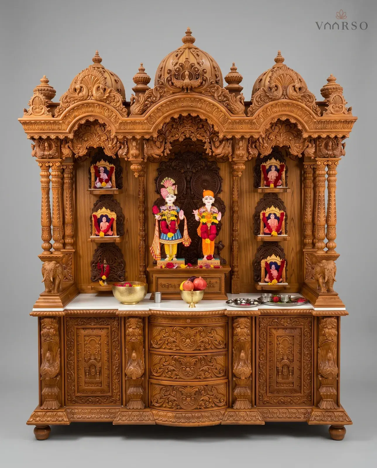 A wide mandir in a warm natural polish, three domes over a carved valance, murtis at the centre with framed figures on six side shelves, brass vessels on a white marble shelf, and curved drawers between two carved cupboards