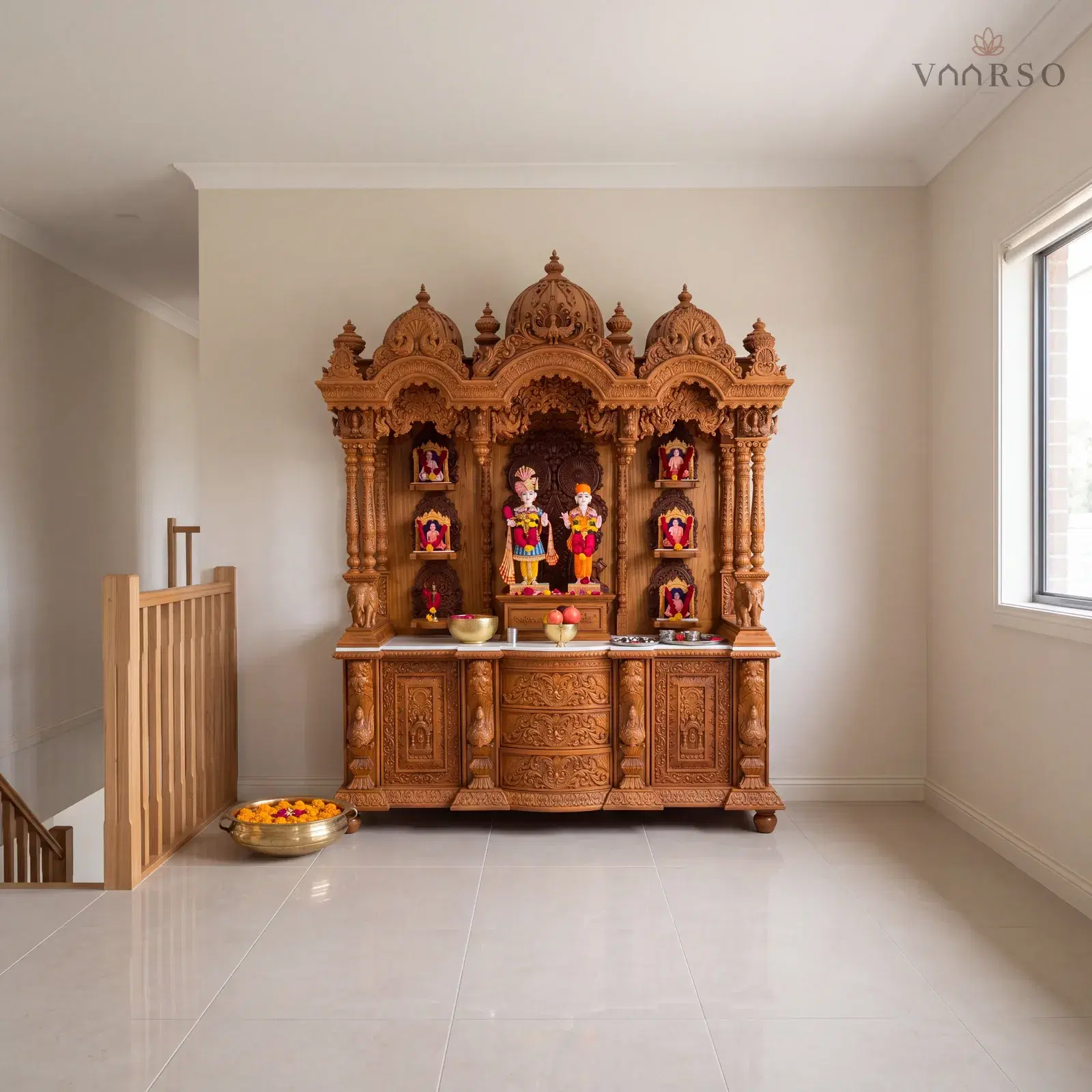 The same mandir in a wide upstairs landing, the stair balustrade along the open side, sand-toned porcelain tile underfoot and a brass urli of floating marigolds on the floor
