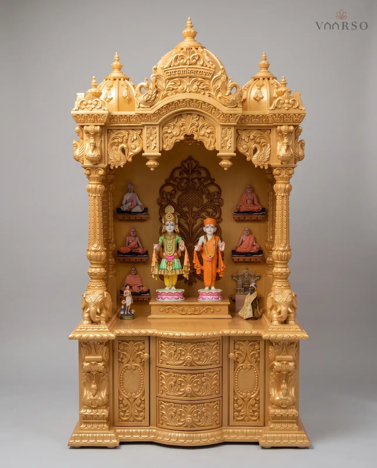 A tall mandir in a pale gold polish, three domes with an inscribed centre over a carved arch, murtis on lotus plinths with seated figures on the side shelves, elephants at the columns, and carved drawers between two cupboards