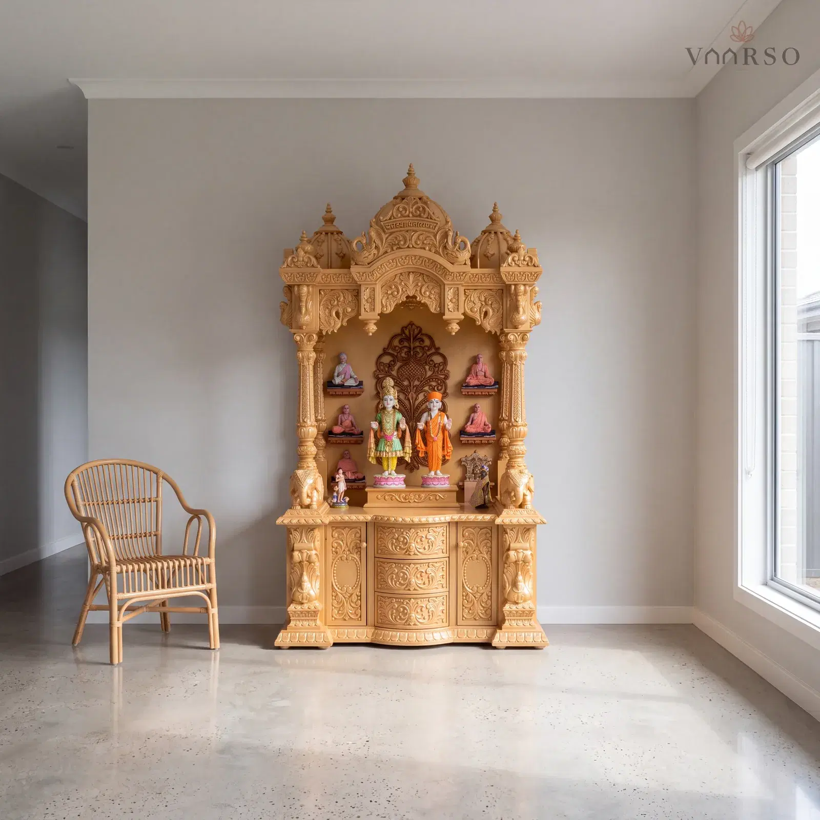 The same mandir in a quiet corner of an open-plan living room, a wide window to one side, polished concrete underfoot and a rattan armchair turned toward it