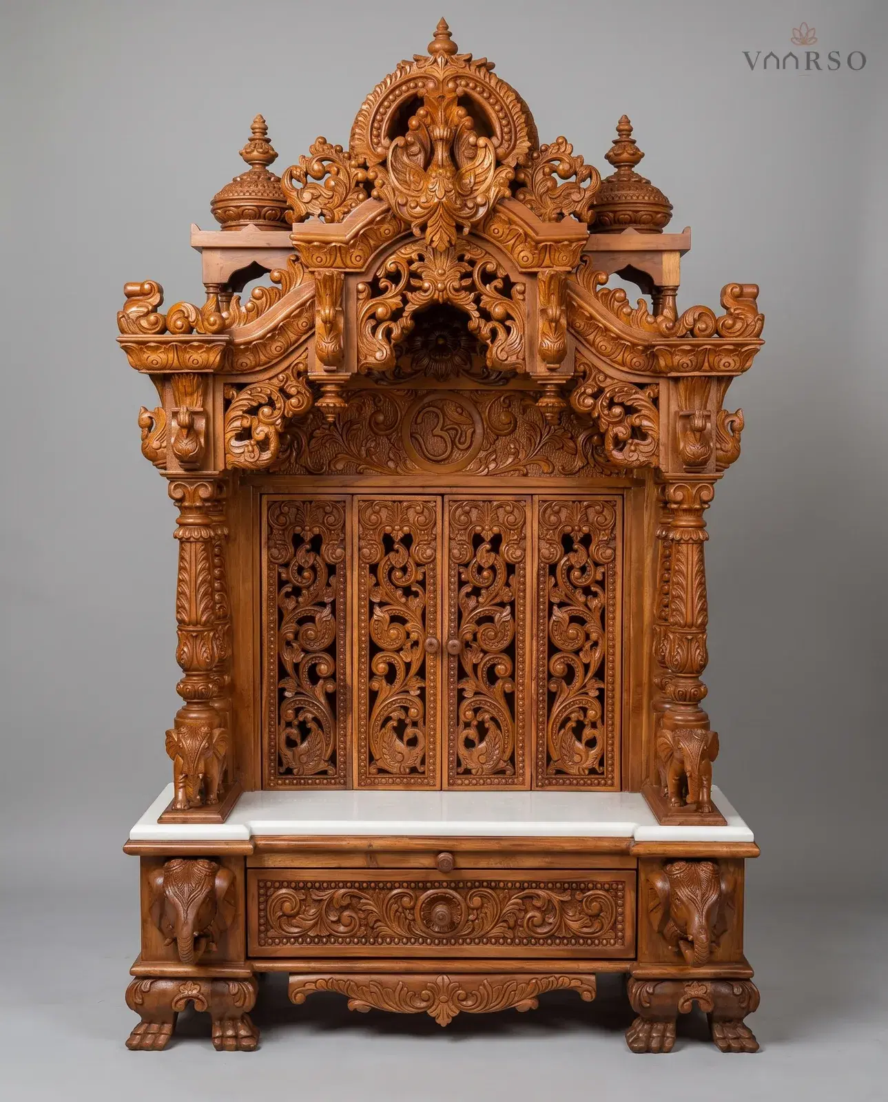 A tall mandir in a warm natural polish, a scrolled crest with an Om panel beneath it, four pierced carved doors closing the shrine, elephants at the columns and elephant heads at the base, over a marble shelf and a single long drawer